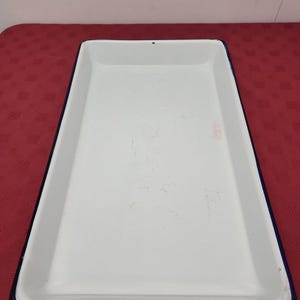 Puede incluir: Una bandeja rectangular de esmalte blanco con un borde azul oscuro. La bandeja presenta una superficie lisa y brillante con sutiles signos de desgaste y ligeros arañazos visibles en la base interior, y una pequeña marca oscura.