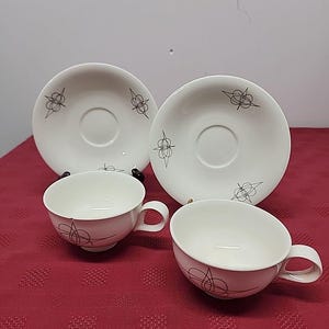 May include: A set of four vintage teacups and saucers. The cups and saucers are white with a black abstract design. The cups have a rounded shape with a handle. The saucers are round with a recessed center.