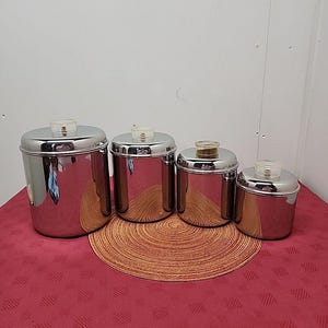 Könnte beinhalten: Ein Set aus vier Edelstahlbehältern mit transparenten Kunststoffdeckeln. Die Behälter sind in einer Reihe angeordnet, wobei die Größe von links nach rechts abnimmt. Sie stehen auf einem runden, gewebten Platzset und einer roten, gemusterten Tischdecke.