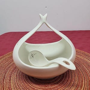 May include: A white ceramic serving bowl with a matching spoon. The bowl has a unique handle design and a small decorative element on the spoon. The bowl and spoon are on a red and brown woven placemat.