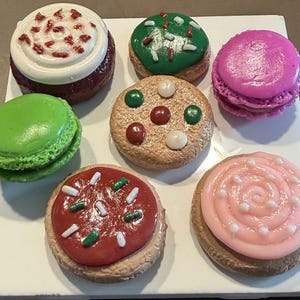 May include: A collection of seven round decorative items resembling assorted cookies and macarons. The set includes a red cookie with white frosting, a green macaron, a green cookie with sprinkles, a tan cookie with colorful dots, a pink macaron, a red cookie with white and green sprinkles, and a tan cookie with pink swirled frosting.