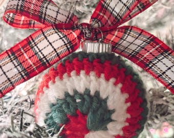 May include: A handmade Christmas ornament with a red, white, and green crocheted design. The ornament is topped with a silver cap and a red and white plaid ribbon bow. The ornament is hanging on a snow-covered Christmas tree.