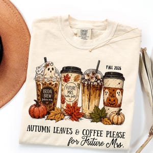May include: A cream-colored t-shirt featuring a graphic of four coffee cups decorated with autumn leaves, pumpkins, and ghosts. The text reads "Bridal Brew", "Future Mrs. hello, fall!", "FALL 2026", and "AUTUMN LEAVES & COFFEE PLEASE for Future Mrs."