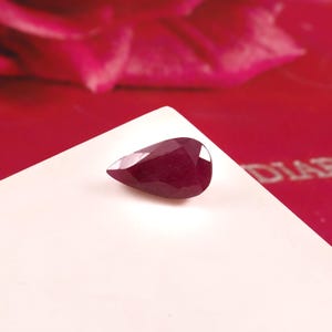 May include: A faceted, pear-shaped ruby gemstone. The deep red gem is set against a white surface, with a blurred red background. The ruby's cut allows light to reflect, creating a sparkling effect. The gemstone is a focal point.