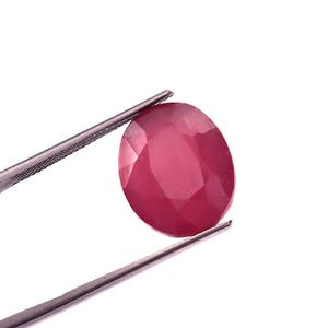 Pode incluir: Uma gema de rubi facetada, em forma oval, segurada por pinças prateadas sobre um fundo branco. O rubi é de uma cor vermelha profunda e rica, com facetas visíveis que capturam a luz. As pinças estão posicionadas para mostrar a gema.