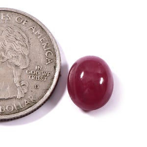 May include: A polished, oval-shaped, deep red gemstone next to a quarter coin. The gemstone has a smooth, glossy surface. The coin is silver and shows the words "In God We Trust" and "Dollar".
