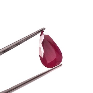 May include: A faceted, teardrop-shaped ruby gemstone held by a pair of silver tweezers. The ruby is a deep, rich red color with multiple reflective facets. The background is plain white.