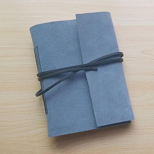 May include: A gray suede journal with a wraparound black leather tie. The journal is closed, with the cover folded over the pages. The pages are visible along the spine. The journal is on a light wood surface.