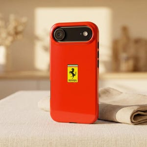 May include: A bright red smartphone case featuring a rectangular Ferrari logo in the center. The logo displays a black prancing horse on a yellow background with a green, white, and red stripe at the top and the word Ferrari printed in black.