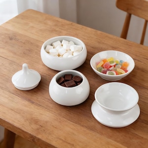 Puede incluir: Conjunto de cuencos de cerámica blanca sobre una mesa de madera. Los cuencos contienen malvaviscos, chocolates y caramelos de colores. También hay un pequeño recipiente con tapa. Ideal para servir aperitivos o postres.