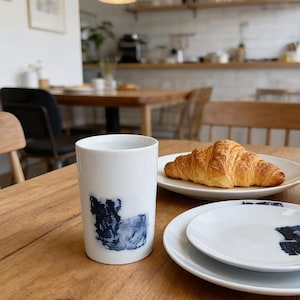 Könnte beinhalten: Ein weißer Keramikbecher mit blauem, abstraktem Design, zusammen mit einem Teller mit einem Croissant und zwei weißen Tellern mit passenden blauen Designs. Die Gegenstände stehen auf einem Holztisch, was auf ein Frühstück hindeutet.
