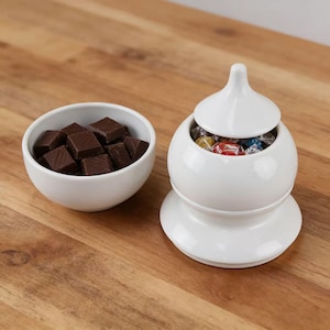 Pode incluir: Uma bomboniere de cerâmica branca com tampa, cheia de doces embalados, fica ao lado de uma pequena tigela branca cheia de chocolates quadrados. Os itens estão em uma superfície de madeira.