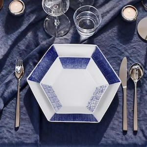 May include: A white and blue hexagonal plate set with a matching bowl, fork, knife, and spoon. The plates have a blue patterned border and a geometric design. The table setting includes water glasses and candles on a blue tablecloth.