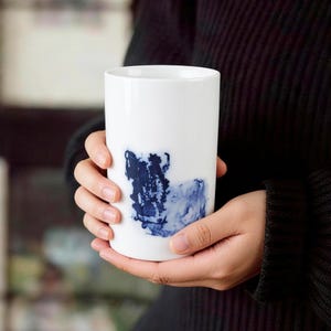 Puede incluir: Una taza de cerámica blanca con forma cilíndrica. La taza presenta un diseño abstracto azul en la parte delantera. La taza está sostenida por una persona que lleva un jersey negro.