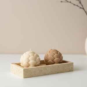Könnte beinhalten: Zwei dekorative Kerzen in Muschelform, eine cremefarbene und eine braune, stehen in einem rechteckigen beigefarbenen Tablett. Die Kerzen haben ein strukturiertes, fächerartiges Design. Eine weiße Vase mit Zweigen befindet sich im Hintergrund.