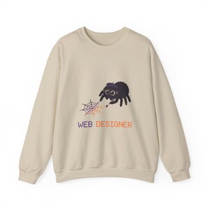 Könnte beinhalten: Beiges Sweatshirt mit einem Cartoon-Spinnen-Design und dem Text "WEB DESIGNER". Die Spinne ist schwarz mit einem lila Hut und hält ein Netz. Der Text ist in Orange und Lila gehalten. Das Sweatshirt hat lange Ärmel.