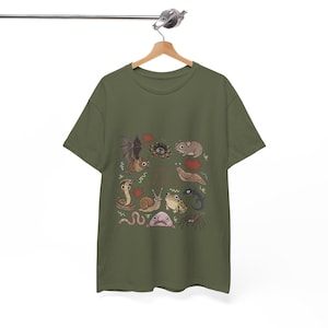 Könnte beinhalten: Ein olivgrünes T-Shirt mit einem zentralen Grafikdruck verschiedener kleiner Tiere und Kreaturen, darunter eine Fledermaus, eine Spinne, eine Maus, eine Nacktschnecke, eine Schlange, eine Schnecke, ein Frosch, eine Eidechse, ein Wurm, ein Blobfisch und eine weitere Spinne.