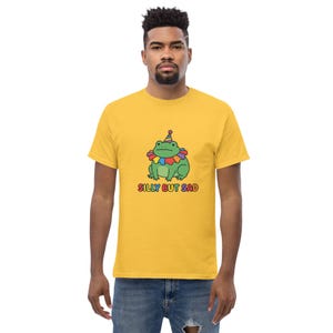 Op de afbeelding: Mosterdgeel T-shirt met een cartoon kikker met een feesthoedje en een kleurrijke gefranjerde kraag. De tekst "SILLY BUT SAD" staat onder de kikker in regenboogkleuren. Het shirt is gemaakt van een zacht materiaal.