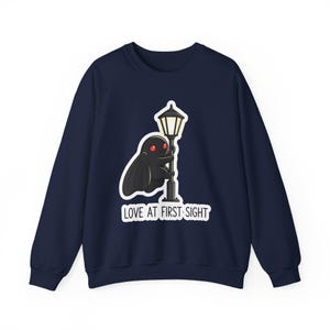 Könnte beinhalten: Marineblaues Sweatshirt mit einer Cartoon-Illustration einer schwarzen Kreatur mit roten Augen, die sich an einer Straßenlaterne festhält. Der Text "LOVE AT FIRST SIGHT" steht darunter. Lässiger Stil.