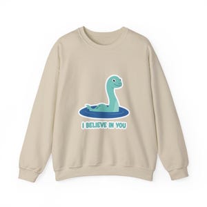 Puede incluir: Sudadera beige con una ilustración de dibujos animados de un monstruo del lago Ness turquesa en agua azul. El texto "I BELIEVE IN YOU" está debajo. La sudadera tiene mangas largas y cuello redondo.