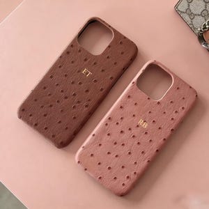 Puede incluir: Dos fundas para teléfono con un patrón texturizado y punteado. Una es marrón oscuro, la otra es rosa empolvado. Cada funda tiene un monograma dorado: "ET" en la funda marrón y "RB" en la funda rosa. Las fundas están sobre una superficie rosa.