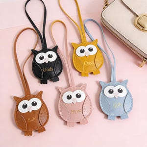 Puede incluir: Una colección de llaveros con forma de búho en varios colores, incluyendo marrón, negro, amarillo, rosa y azul. Cada llavero presenta un diseño cosido con ojos blancos y una correa de cuero. Algunos llaveros tienen texto: "Gnds", "Oasx", "Byue", "Ois" y "Sahl".