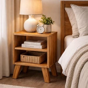 May include: A wooden nightstand with two shelves, a lamp, and a small alarm clock. The lamp has a white, ribbed base and a beige lampshade. A small potted plant and a woven basket are also on the nightstand. The bed has a white comforter and a beige blanket.
