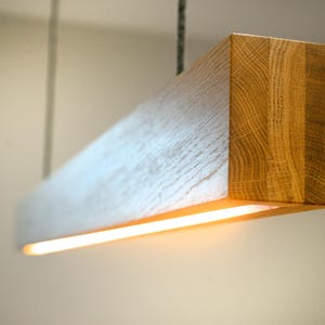 May include: A rectangular wooden pendant light. The end of the light has a natural wood grain finish, while the sides are white-washed. A warm, yellow light shines from the bottom. The light hangs from a chain.