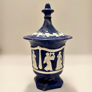 May include: A blue and white ceramic jar with a lid. The jar features a decorative design of figures in white relief against a blue background. The lid has a white floral pattern. The jar is approximately 8 inches tall.