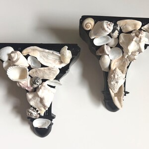 May include: Two black decorative wall sconces, each adorned with a variety of seashells in shades of white, cream, and tan. The shells are arranged on the sconces, creating a coastal or nautical theme. The sconces are mounted on a white wall.