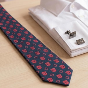 Puede incluir: Una corbata azul marino con un patrón geométrico rojo repetido y gemelos a juego. La corbata está junto a una camisa blanca con los gemelos apoyados en el puño. Los gemelos tienen un diseño a juego.