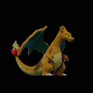 Könnte beinhalten: Gestrickte Charizard-Figur in Orange-, Gelb- und Grüntönen. Der Drache hat detaillierte Flügel, eine Flamme am Schwanz und weiße Akzente an den Füßen. Die Figur ist vor schwarzem Hintergrund.