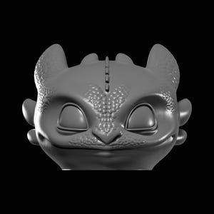 Op de afbeelding: Een grijs, 3D-geprint model van een drakenkop, met gesloten ogen en een zachte glimlach. De draak heeft een getextureerde huid, kleine oren en een rij stekels langs de bovenkant van zijn hoofd. De achtergrond is effen zwart.