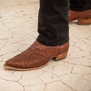 Könnte beinhalten: Braune Cowboy-Stiefel mit einem strukturierten, schuppenartigen Muster. Die Stiefel haben eine spitze Zehenpartie und einen gestapelten Absatz. Die Person trägt eine schwarze Hose. Die Stiefel sind ein klassischer Western-Stil.