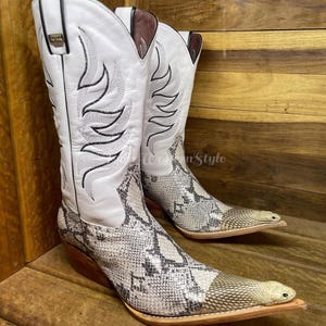 Può includere: Un paio di stivali da cowboy in pelle bianca con cuciture nere e un motivo a pelle di serpente sulla parte inferiore dello stivale. Gli stivali hanno la punta a punta e un tacco in legno marrone. Gli stivali sono esposti su una superficie di legno.