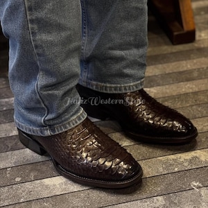 Könnte beinhalten: Braune Leder-Cowboystiefel mit einem strukturierten, schuppenartigen Muster, getragen mit blauen Jeans. Die Stiefel haben eine spitze Zehenpartie und einen niedrigen Absatz. Die Stiefel stehen auf einem grau-braun gestreiften Boden.