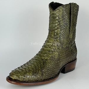 Può includere: Un singolo stivale da cowboy verde oliva con punta e tacco impilato. Lo stivale presenta un motivo a pelle di serpente e una suola marrone scuro. Lo stivale è realizzato in pelle e ha un classico design western.