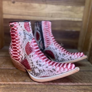 May include: A pair of ankle boots with a pointed toe and a stacked heel. The boots feature a snakeskin pattern in shades of red, pink, and white. The boots have a Western style and are made of leather.