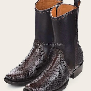 Könnte beinhalten: Ein Paar dunkelbraune und schwarze Leder-Cowboystiefel. Die Stiefel haben eine quadratische Spitze und ein strukturiertes, schlangenartiges Muster auf dem unteren Teil. Der obere Schaft ist aus glattem schwarzem Leder. Die Stiefel haben eine Zuglasche und einen Reißverschluss.