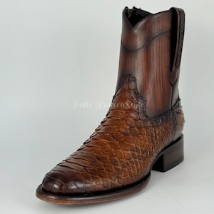 Könnte beinhalten: Ein brauner Leder-Cowboy-Stiefel mit einem strukturierten Schlangenhautmuster am Fuß und einem glatten, dunkelbraunen Schaft. Der Stiefel hat eine quadratische Spitze und einen kurzen Absatz. Der Stiefel ist mittelbraun.