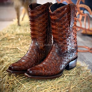 Puede incluir: Un par de botas de vaquero de cuero marrón con un patrón texturizado en forma de escamas. Las botas tienen una punta puntiaguda, un tacón apilado y una caña alta. Las botas se exhiben sobre un lecho de heno.