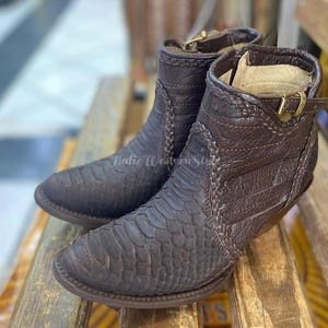 Könnte beinhalten: Ein Paar braune Leder-Ankle Boots mit einem strukturierten, schuppenartigen Muster. Die Stiefel haben eine spitze Zehenpartie, einen kleinen Absatz und eine Schnalle. Die Stiefel stehen auf einer Holzoberfläche.