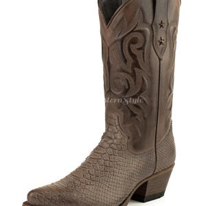 Può includere: Stivale da cowboy in pelle marrone con punta affilata e motivo a pelle di serpente strutturato sul piede. Lo stelo superiore presenta cuciture decorative e due piccoli accenti a stella. Lo stivale ha un tacco impilato.