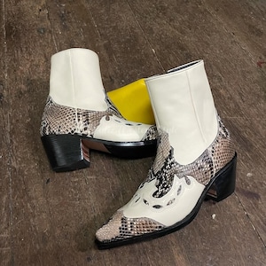 Könnte beinhalten: Ein Paar cremefarbene Cowboy-Stiefel mit Schlangenmuster an den Seiten und der Spitze. Die Stiefel haben eine spitze Zehenpartie, einen schwarzen Absatz und dekorative Aussparungen am Blatt. Der Schaft ist einfarbig cremefarben.