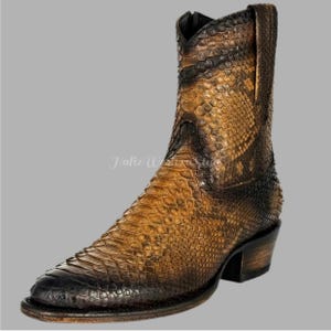 Könnte beinhalten: Ein brauner und schwarzer Cowboy-Stiefel mit spitzer Zehenpartie. Der Stiefel besteht aus einem schlangenlederartigen Material mit einem strukturierten Muster. Der Stiefel hat einen kurzen Schaft und einen kleinen Absatz. Der Text "Hafiz Western Style" ist sichtbar.