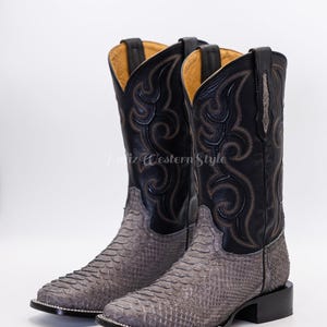 Peut inclure: Paire de bottes de cowboy avec un motif peau de serpent gris sur le pied et une tige en cuir noir. Les bottes ont des coutures décoratives et un bout carré. La tige est noire avec des coutures marron.