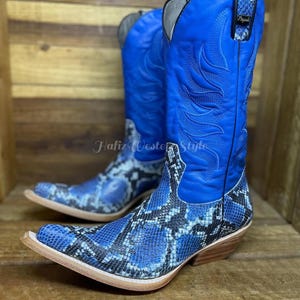 May include: A pair of blue cowboy boots with a snakeskin pattern on the foot and ankle. The shaft is solid blue with decorative stitching. The boots have a pointed toe and a stacked heel. The text "Hafiz Western Style" is visible.