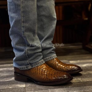 Puede incluir: Botas de vaquero de cuero marrón con un patrón texturizado similar al de un reptil. Las botas tienen una punta puntiaguda y un tacón bajo. Se usan con vaqueros azules. El texto "Hafiz Western Style" es visible.