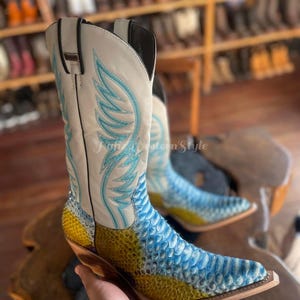 May include: A pair of cowboy boots with a cream-colored shaft featuring turquoise wing embroidery. The lower portion of the boots is patterned with blue and yellow scales. The boots have a pointed toe and a wooden heel. The text "Natural Western Style" is visible.
