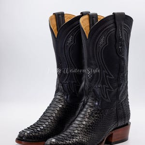 May include: A pair of black cowboy boots with a textured snakeskin pattern on the foot and smooth leather uppers. The boots have decorative stitching and a stacked heel. The interior is a light tan color.